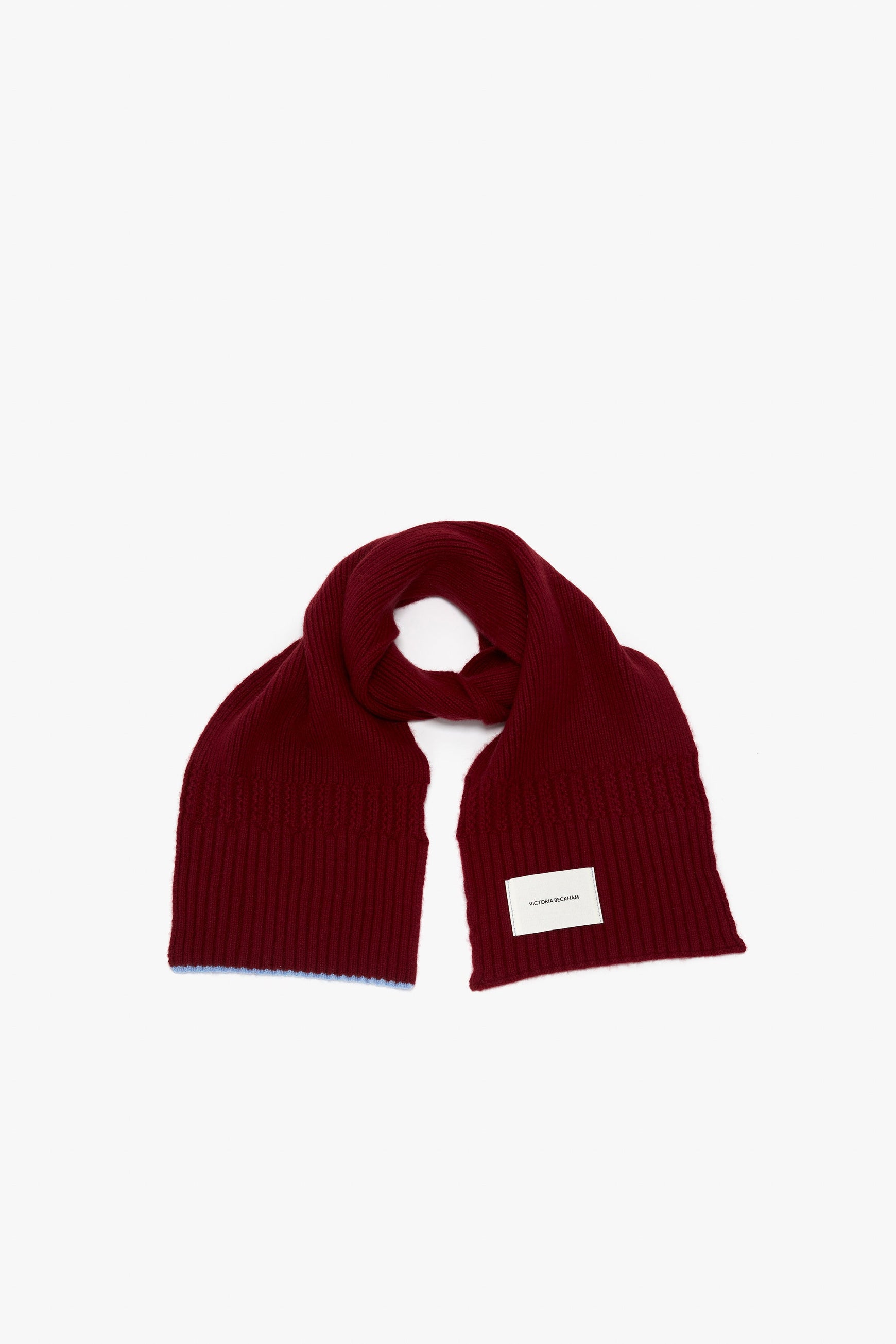 A folded, dark red ribbed scarf with a white rectangular label attached, displayed on a plain white background. The Victoria Beckham Exclusive Logo Patch Scarf In Burgundy channels Victoria Beckham's signature elegance, featuring the iconic logo patch scarf detail that adds a touch of sophistication.