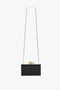 A rectangular black Frame Flower Minaudiere Bag in Black by Victoria Beckham with a long gold chain, a decorative flower clasp, and a detachable shoulder strap.