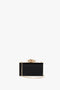 A black rectangular Frame Flower Minaudiere Bag in Black with a gold chain strap and a decorative gold flower clasp on top by Victoria Beckham.