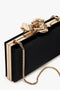 Frame Flower Minaudiere Bag in Black with gold trim and a gold rose flower clasp on top, accompanied by a detachable gold chain shoulder strap by Victoria Beckham.