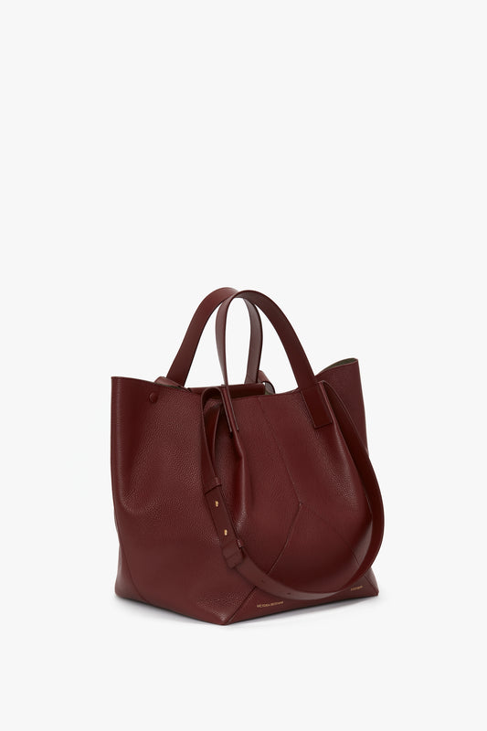 A W11 Medium Tote Bag In Burgundy Leather by Victoria Beckham, featuring two handles and a shoulder strap, displayed on a plain white background. Its versatile styling makes it perfect for any occasion.