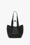 A W11 Medium Tote Bag In Black Leather with dual shoulder straps, an open top, and a textured surface, set against a plain white background. This Victoria Beckham creation showcases the elegance of premium leather goods.