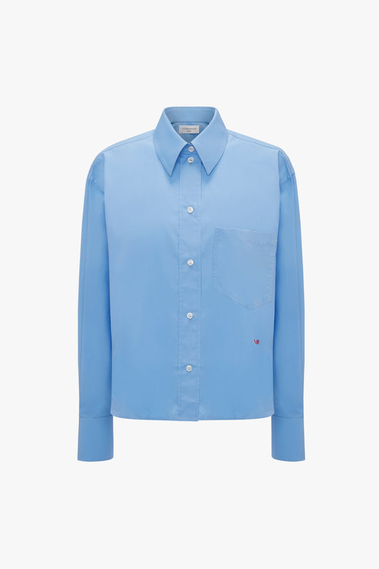 A Cropped Long Sleeve Shirt In Oxford Blue by Victoria Beckham with a single chest pocket and a small embroidered heart near the bottom right, made from organic cotton for a relaxed fit.