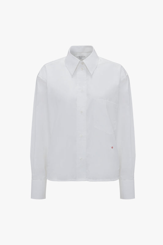 A Cropped Long Sleeve Shirt In White by Victoria Beckham with a pointed collar and a single chest pocket, displaying a small "WD" monogram. This piece features relaxed tailoring for ultimate comfort.