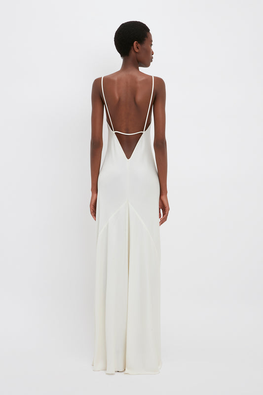 A person stands facing away, wearing a long, backless white Victoria Beckham Floor-Length Cami Dress In Ivory made of crepe back satin with thin straps. The dress has a V-shaped detail on the back and flows down to the floor, evoking timeless 90s fashion.
