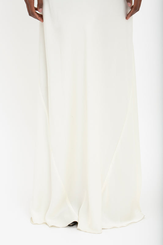 A person wearing a long, white Floor-Length Cami Dress In Ivory by Victoria Beckham stands against a white background, their lower half reminiscent of 90s fashion elegance in crepe back satin.