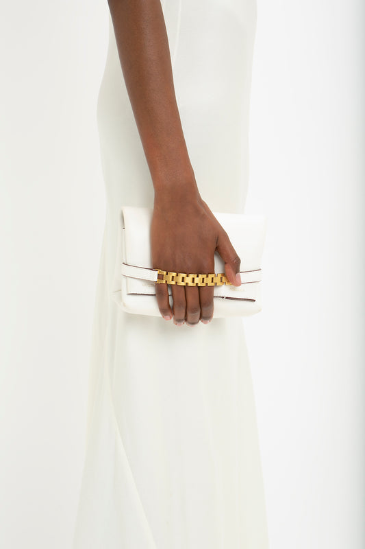 A person in a Floor-Length Cami Dress In Ivory by Victoria Beckham holds a white clutch bag with a gold chain strap in their left hand, channeling 90s fashion vibes.