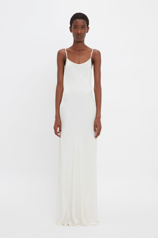 A person stands facing forward, wearing the Victoria Beckham Floor-Length Cami Dress In Ivory. The background is plain white, evoking a touch of 90s fashion.