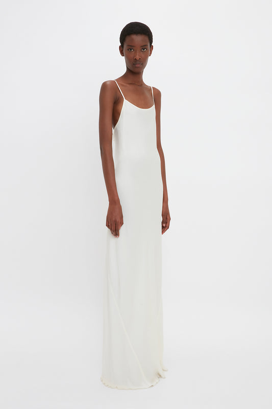A person stands against a white background wearing the Floor-Length Cami Dress in Ivory by Victoria Beckham. The crepe back satin fabric drapes elegantly over their figure. They have short hair and a neutral expression.