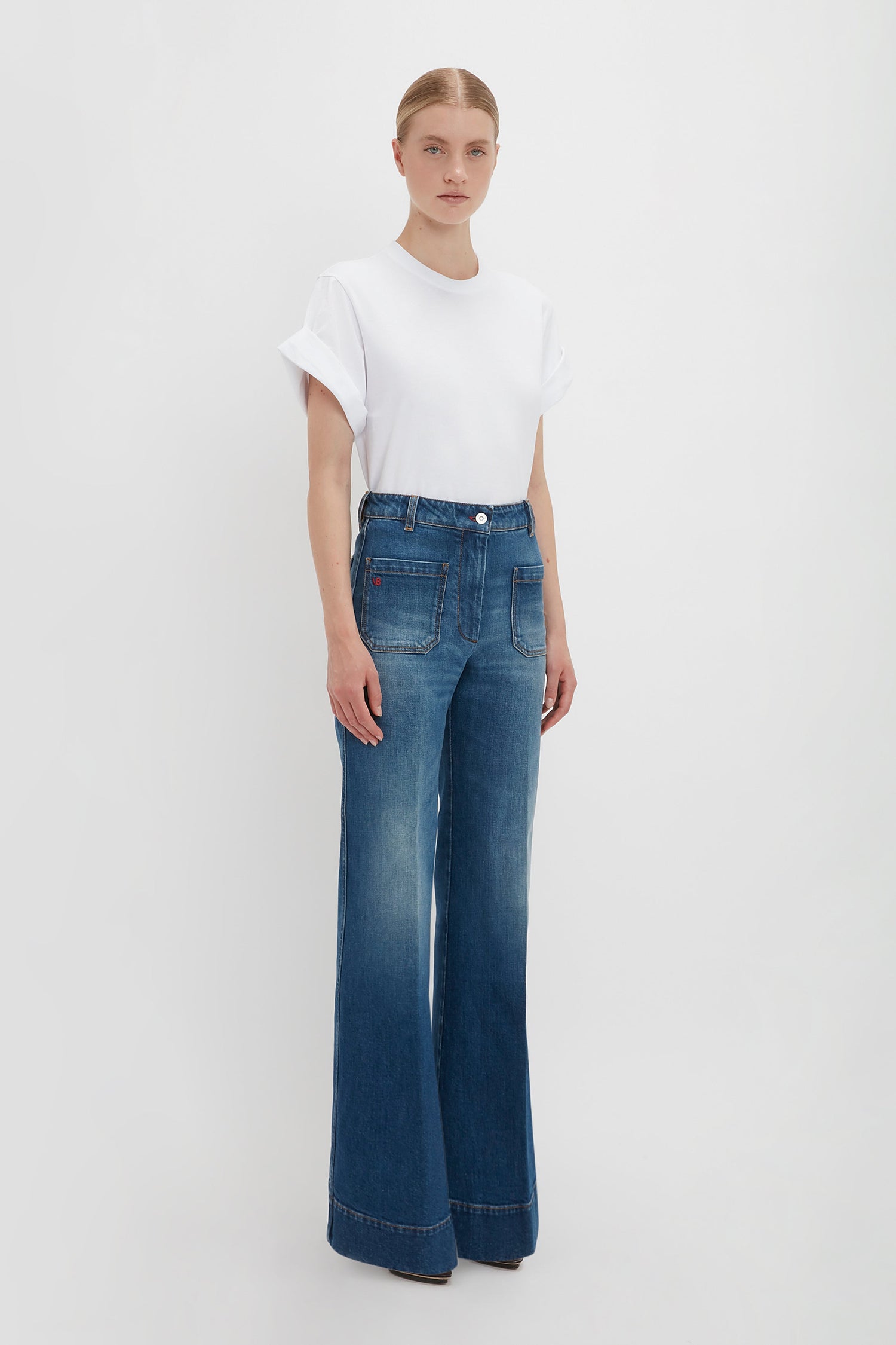 Woman in a Victoria Beckham Asymmetric Relaxed Fit T-Shirt in White and blue high-waisted jeans standing against a white background.