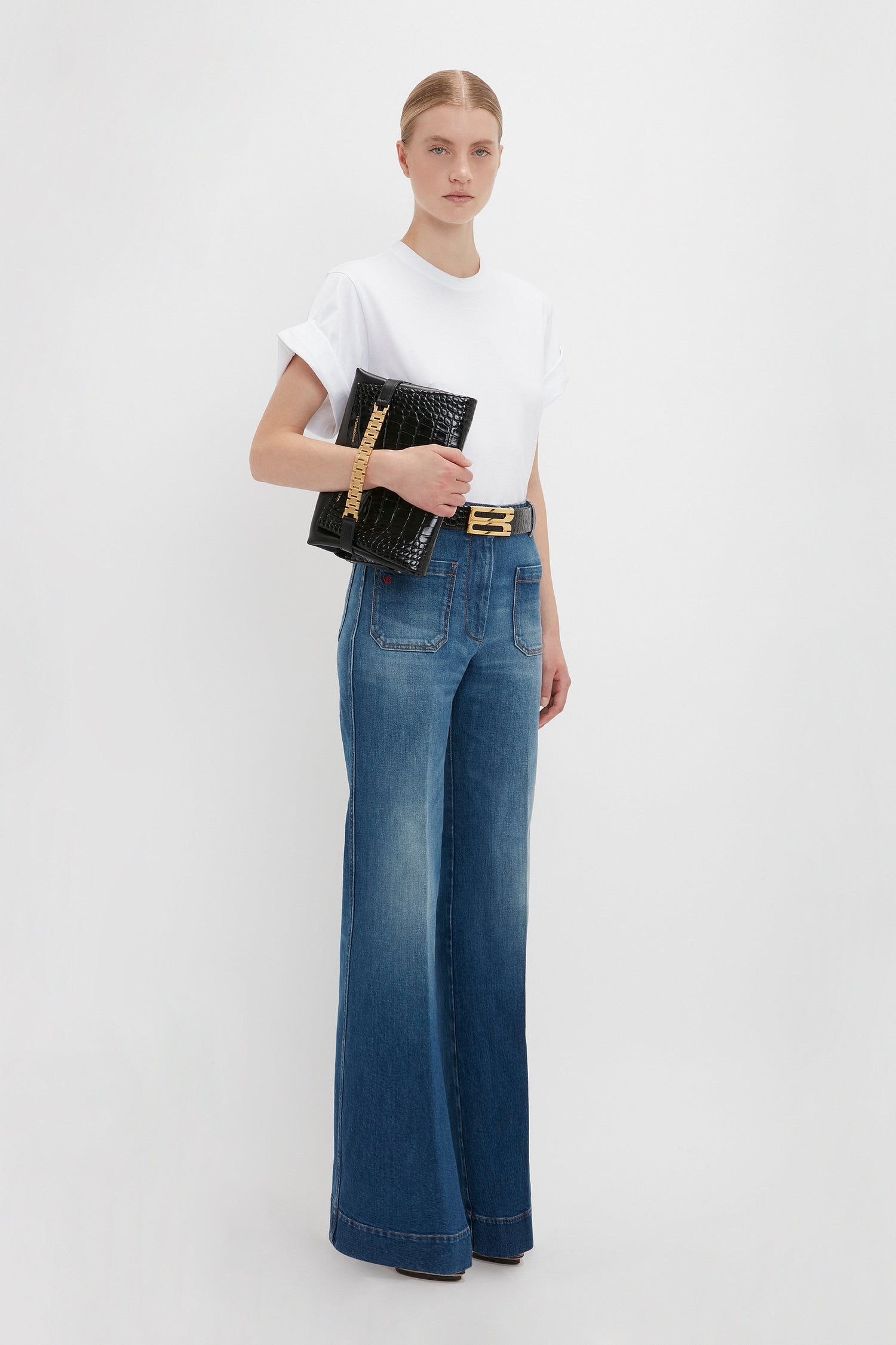 A woman in an oversized Victoria Beckham Asymmetric Relaxed Fit T-Shirt in white and blue jeans holding a black clutch and wearing a gold belt against a white background.