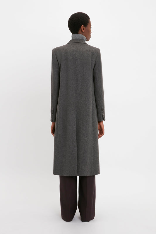 Person wearing a long, gray "Tailored Slim Coat In Grey Melange" by Victoria Beckham and dark pants seen from the back, standing against a plain white background, showcasing a flattering silhouette.
