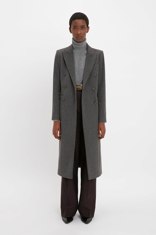 A person stands facing the camera, wearing a Victoria Beckham Tailored Slim Coat In Grey Melange over a gray turtleneck and dark trousers, against a plain white background. The ensemble's structured design draws from military menswear, creating a flattering silhouette.