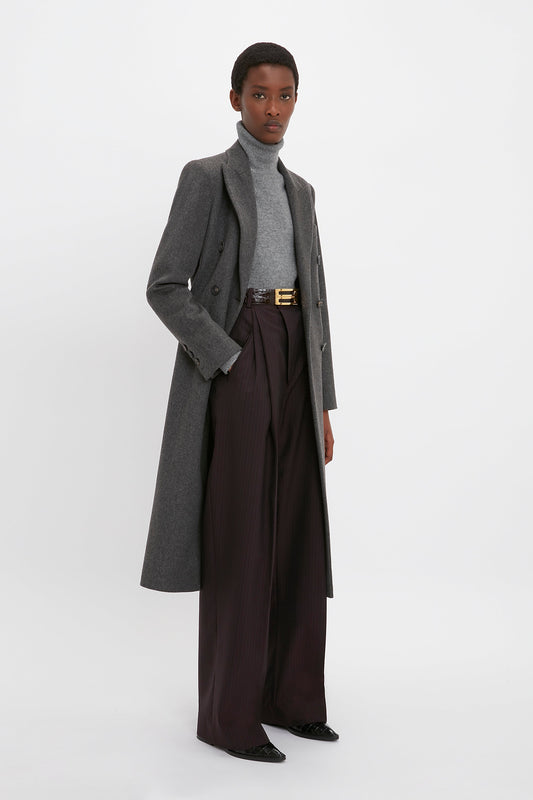 Person wearing a gray turtleneck, Victoria Beckham Tailored Slim Coat In Grey Melange, and high-waisted dark pleated trousers, standing against a white background.