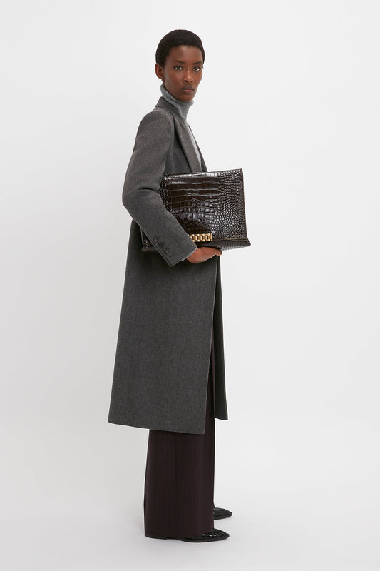 A person in a Victoria Beckham Tailored Slim Coat In Grey Melange and dark pants stands against a white background, holding a large, dark crocodile-patterned handbag.