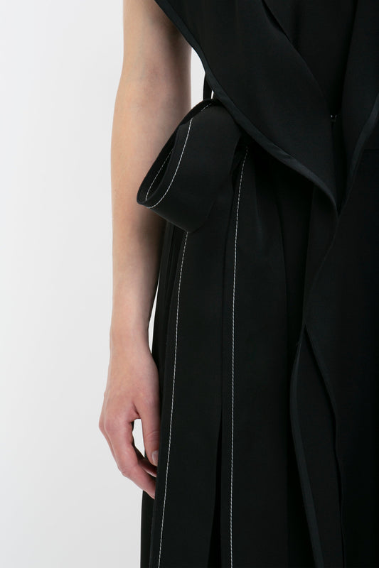 A close-up of a person wearing a wrap-front Trench Dress In Black by Victoria Beckham with white stitching, featuring oversized front flap pockets and a sash-style belt.
