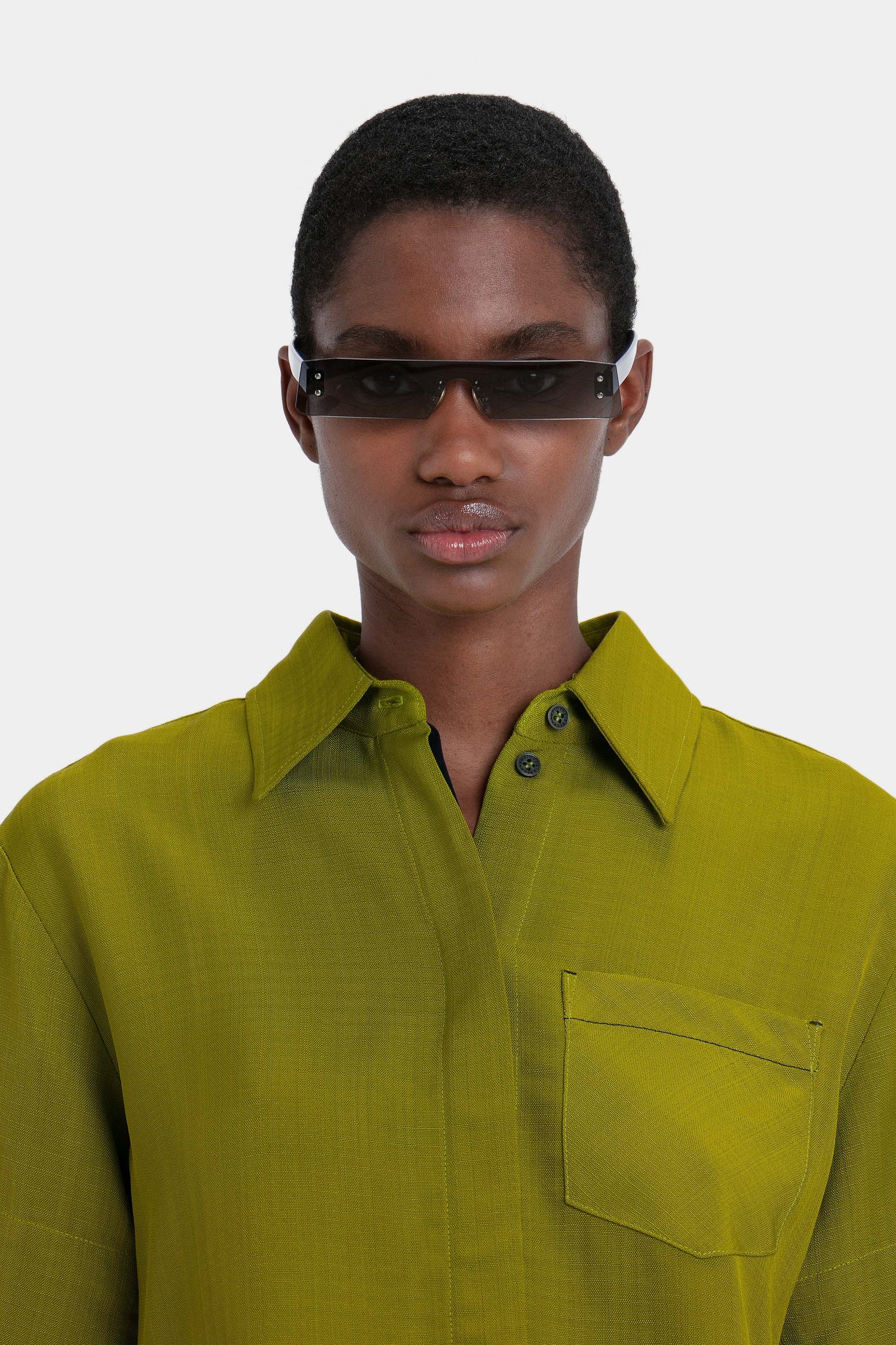 A person wearing Mini Visor Sunglasses In Black-Green and a bright green collared shirt, reminiscent of the SS23 Victoria Beckham collection, stands against a plain background.