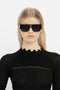 A woman with blonde hair wearing Victoria Beckham V Plaque Frame Sunglasses In Black with a double bridge and a textured black top, standing against a white background.