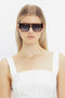 A person with long blonde hair wears statement Victoria Beckham V Plaque Frame Sunglasses In Dark Brown Horn and a white sleeveless top, posing against a plain white background.