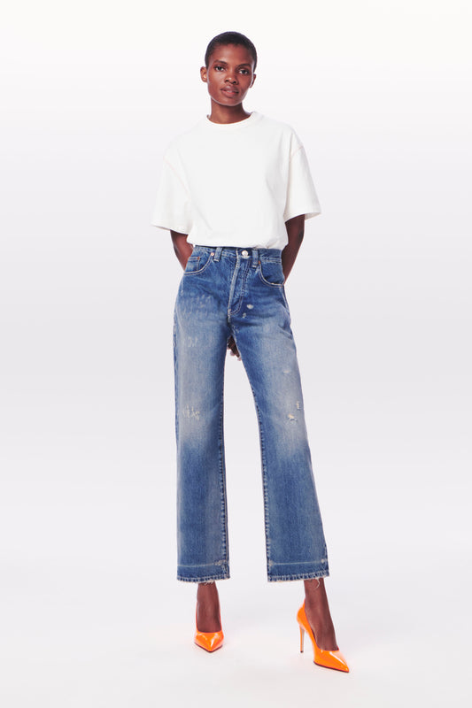 Person stands against a plain background wearing a white T-shirt, Victoria Beckham Victoria Mid-Rise Jean in Vintage Wash, and bright orange high heels.