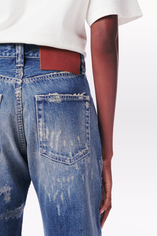 Close-up of a person wearing Victoria Beckham Victoria Mid-Rise Jean in Vintage Wash with a brown leather patch on the waistband, paired with a white t-shirt. The mid-rise jeans have visible wear and tear patterns.