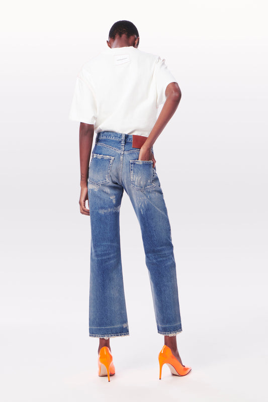 A person wearing a white shirt, Victoria Beckham Victoria Mid-Rise Jean in Vintage Wash, and orange high heels is standing and facing away from the camera.