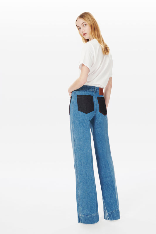 A person with long hair is standing sideways, wearing a white short-sleeve shirt and Victoria Beckham's Alina High Waisted Patch Pocket Jean In 70s Wash.