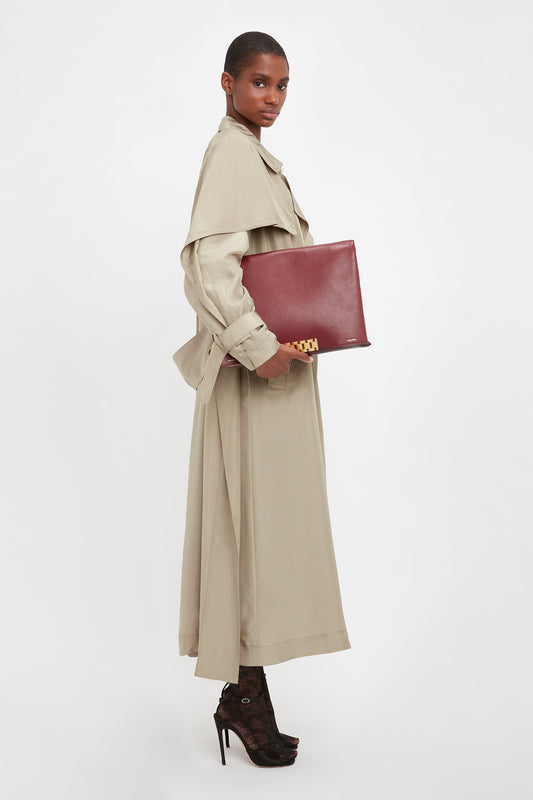 Person standing in profile wearing a long beige trench coat, holding a large red book or folder, and wearing high-heeled shoes. They also have a Victoria Beckham Jumbo Chain Pouch Bag In Bordeaux with a detachable strap made from grained leather. The background is plain white.