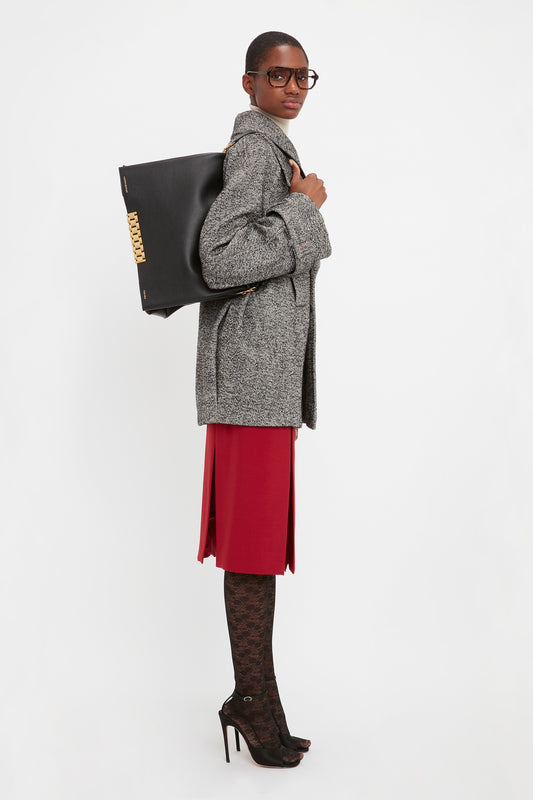 Person wearing glasses, a gray coat, red skirt, patterned tights, and high heels, holding a Jumbo Chain Pouch Bag In Black Leather by Victoria Beckham over their shoulder, standing against a plain white background.