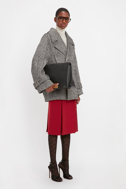 A person wearing a gray coat over a white turtleneck, red skirt, patterned tights, and black high heels, holds a large Victoria Beckham Jumbo Chain Pouch Bag In Black Leather and wears large glasses.