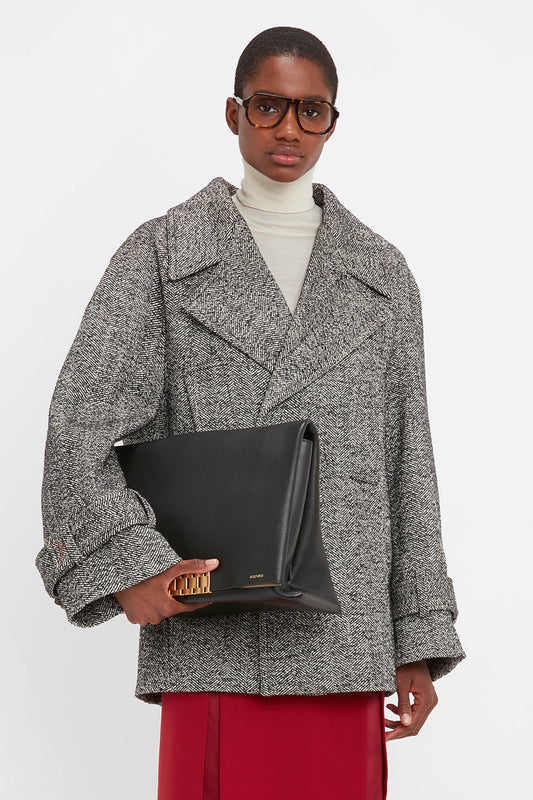 A person wearing a gray coat, turtleneck, and glasses holds a Victoria Beckham Jumbo Chain Pouch Bag In Black Leather against a plain white background.
