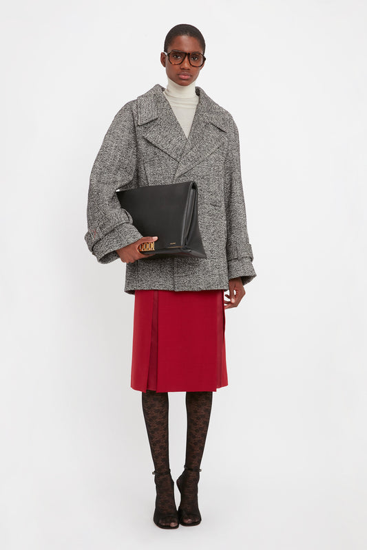 A person stands against a white background wearing a gray oversized coat, a turtleneck, a red skirt, patterned tights, and black heels. They hold a Jumbo Chain Pouch Bag In Black Leather by Victoria Beckham while sporting large glasses.