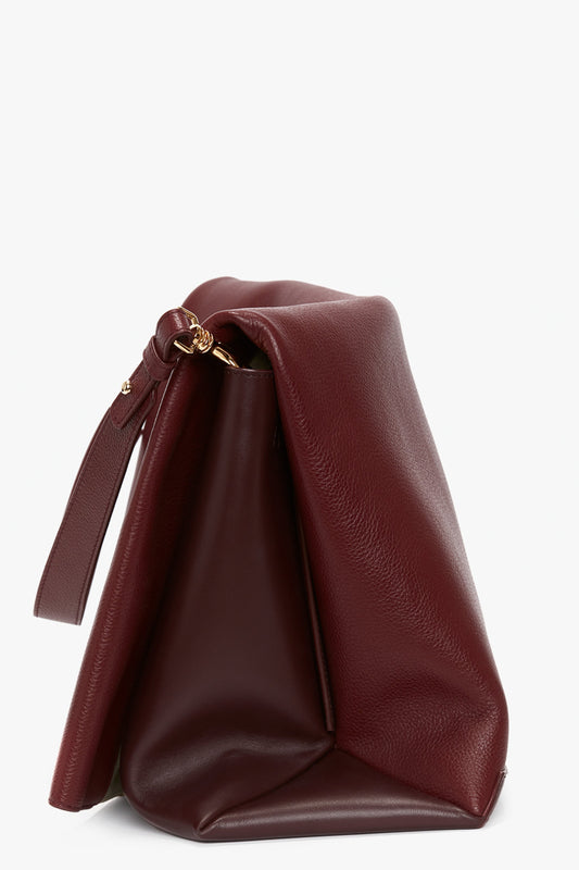 A Victoria Beckham Jumbo Chain Pouch Bag In Bordeaux with a gold clasp and a wrist strap, partially open, showing pleated sides and a textured surface. Featuring grained leather, this elegant piece also comes with a detachable strap for added versatility.
