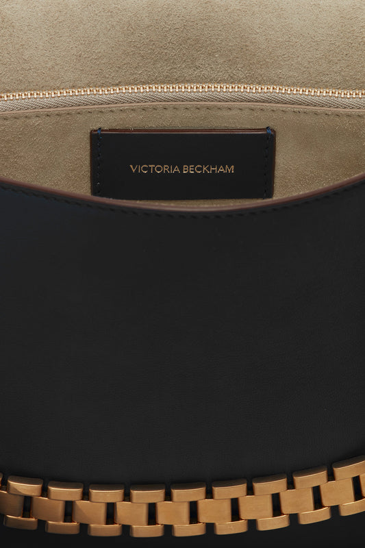 A close-up of an open black Chain Pouch Bag with Strap In Black Leather made of Nappa leather with an interior label reading "Victoria Beckham." The inside is lined with beige material, and the bag features a gold-tone chain.