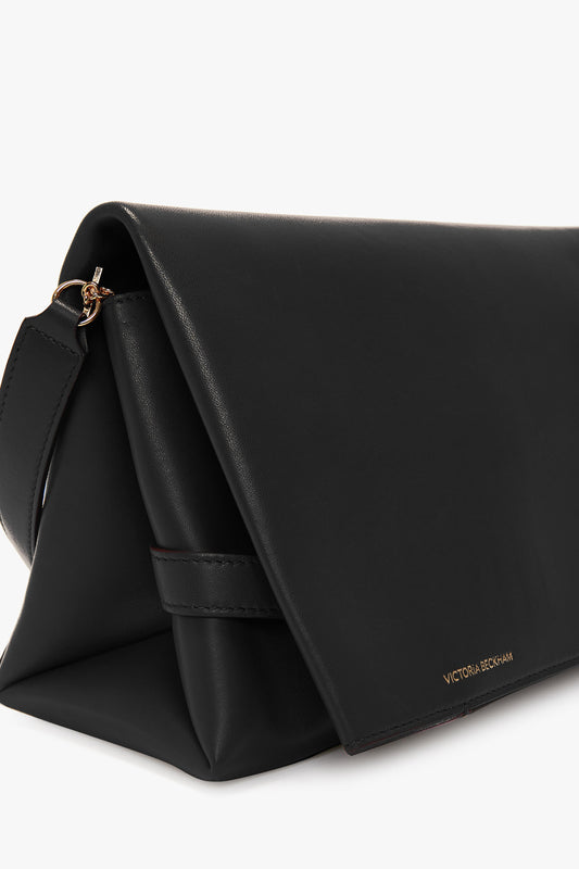 A Chain Pouch Bag with Strap In Black Leather with a folded triangular design, gold-tone chain, and a gold clasp. The name "Victoria Beckham" is printed in gold on the bottom right corner.