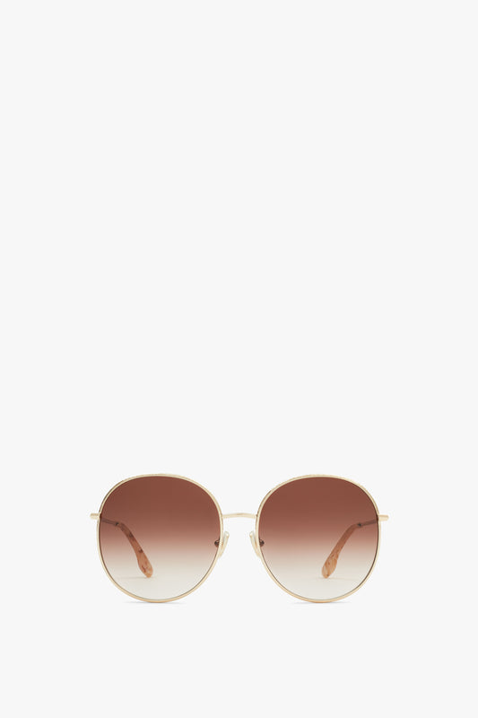 Round, gradient brown-lensed sunglasses with thin gold metal frames and brown temple tips, exuding a vintage-inspired charm against a plain white background. Introducing the Hammered Round Sunglasses In Gold Brown by Victoria Beckham.