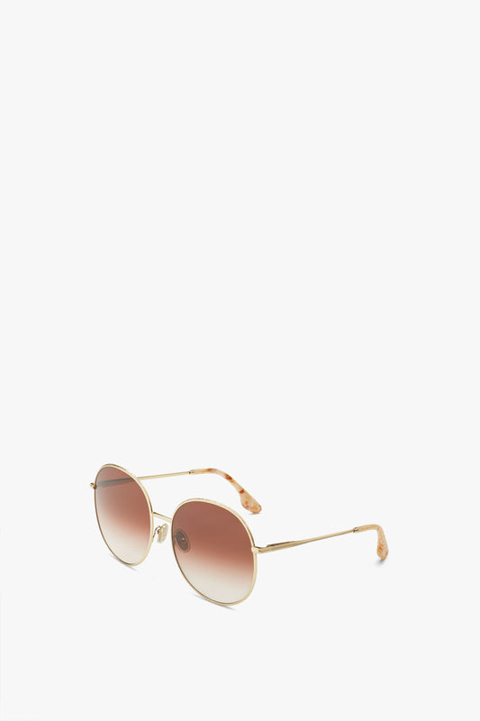 A pair of Victoria Beckham Hammered Round Sunglasses In Gold Brown with vintage-inspired, gold metal frames and brown gradient lenses is displayed against a plain white background.
