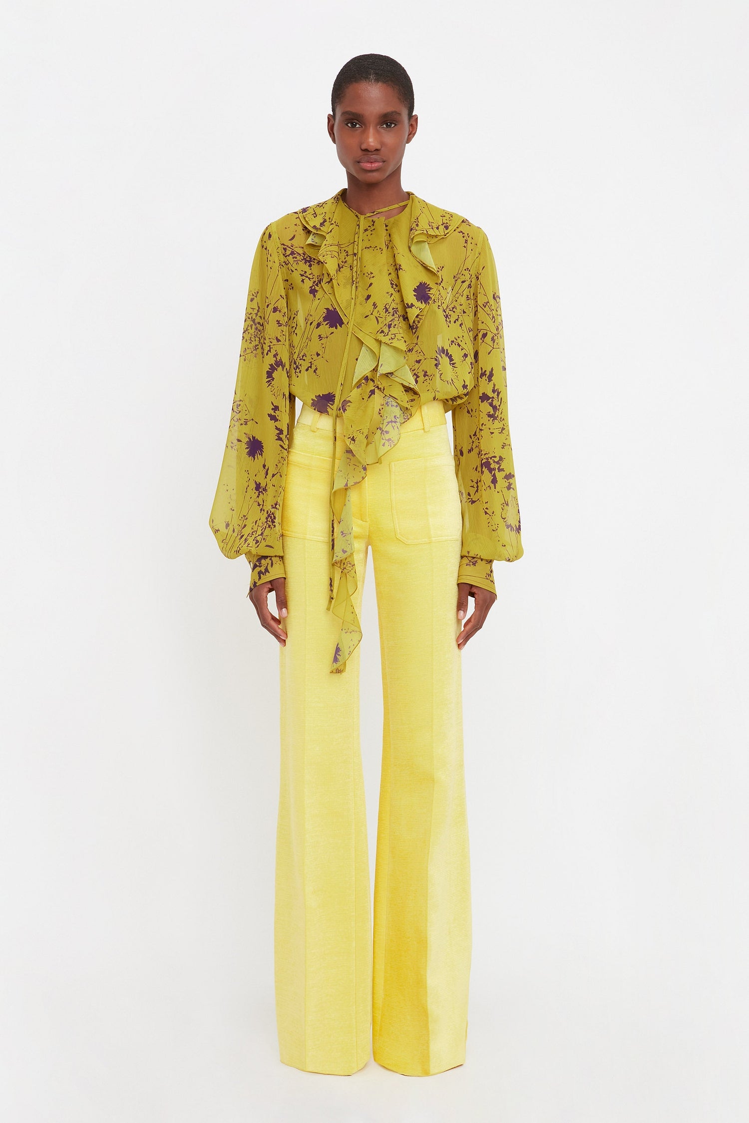 Model wears the Alina tailored trousers in a bright sunflower yellow. These bright yellow wide leg trousers have front detail pockets and a flared hem, made from durable recycled polyester.