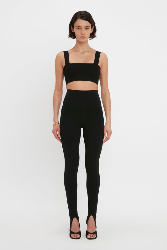 A person wearing a black VB Body Strap Bandeau Top In Black by Victoria Beckham and matching high-waisted leggings stands in front of a plain white background. They are also wearing black heels and a gold bracelet, giving the outfit a contemporary finish.