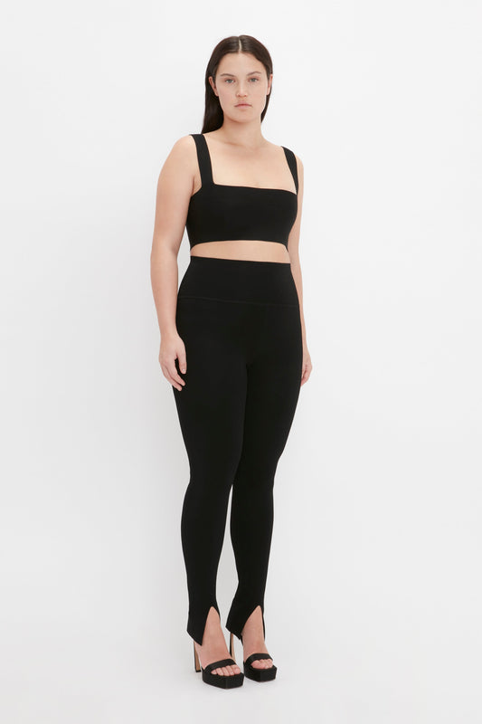 A woman stands against a plain white background wearing a Victoria Beckham VB Body Strap Bandeau Top In Black, ultra form-fitting knitwear high-waisted black leggings, and black high-heeled sandals, achieving a contemporary finish.