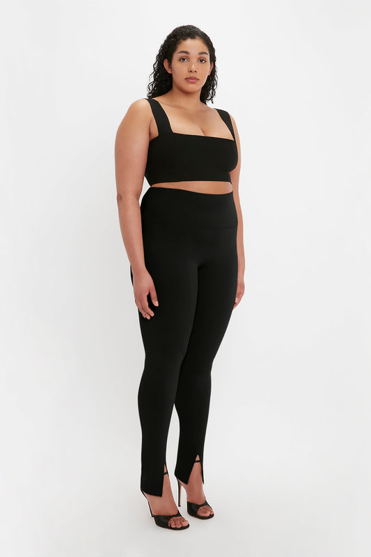 A person with curly hair is wearing an ultra form-fitting knitwear ensemble, featuring a black two-piece outfit with a VB Body Strap Bandeau Top In Black by Victoria Beckham and high-waisted leggings with a slit at the bottom, paired with black heels.