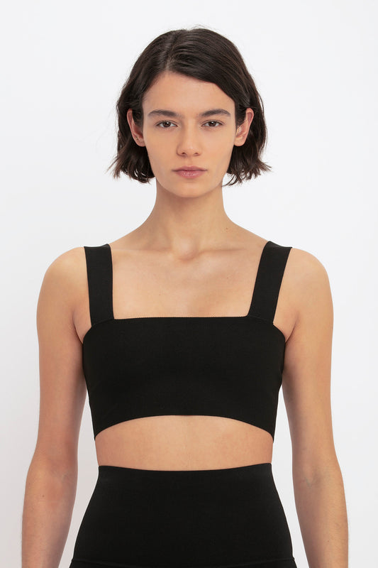 A person with short hair stands against a white background, wearing an ultra form-fitting VB Body Strap Bandeau Top In Black by Victoria Beckham and high-waisted bottoms.