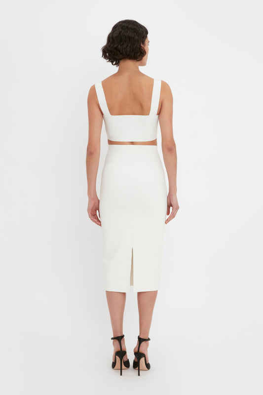 A woman viewed from behind wearing a white sleeveless dress with a square neckline and back, styled with black high heels and a Victoria Beckham Body Strap Bandeau Top in White Midi Skirt.