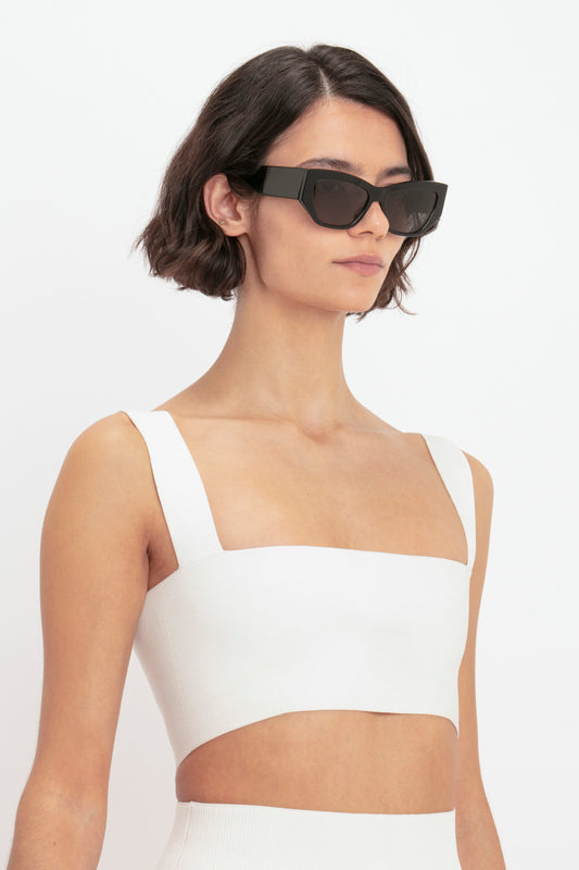 A woman in a white crop top and oversized black sunglasses poses against a plain background, complemented by a Victoria Beckham Body Strap Bandeau Top In White.