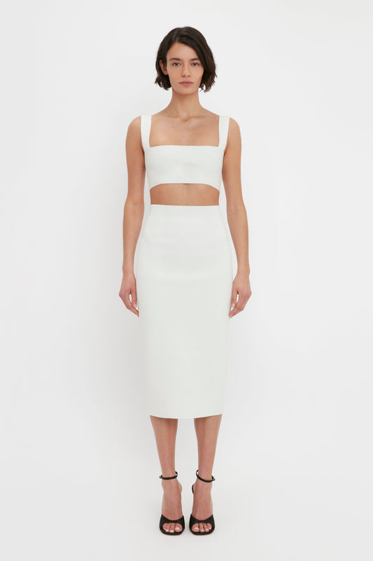 A woman stands against a plain white background, wearing a white two-piece outfit of a Victoria Beckham VB Body Strap Bandeau Top in White and high-waisted skirt. She has short dark hair and is wearing black high-heeled sandals.
