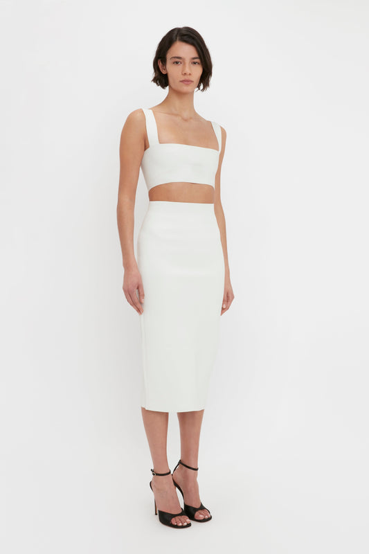 A woman wearing a white two-piece dress with a Victoria Beckham Body Strap Bandeau Top in White and a VB Body fitted midi skirt, paired with black ankle-strap heels, stands against a plain white background.
