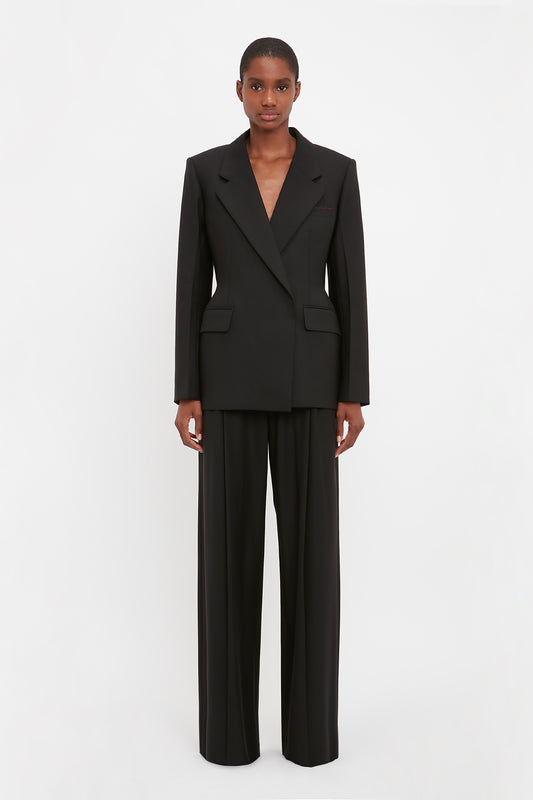 A person stands against a white background wearing the Victoria Beckham Waist Fitted Jacket In Black and matching wide-leg trousers, exuding modern sophistication.