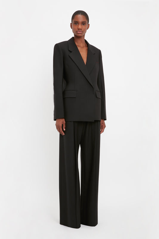 Person standing against a white background, wearing a **Victoria Beckham Waist Fitted Jacket In Black** with matching wide-leg pants, exuding modern sophistication. A subtle contrasting red tailor’s tack adds a touch of distinct elegance to the outfit.