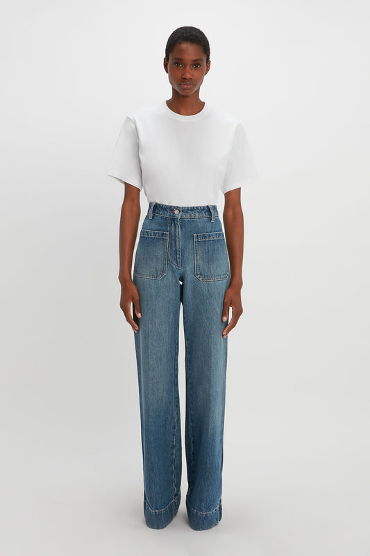 A person stands against a plain white background, wearing a white T-shirt and Victoria Beckham Alina High Waisted Jean In Shadow Wash with high-waisted blue denim silhouette and large front pockets. They appear to be looking directly at the camera.