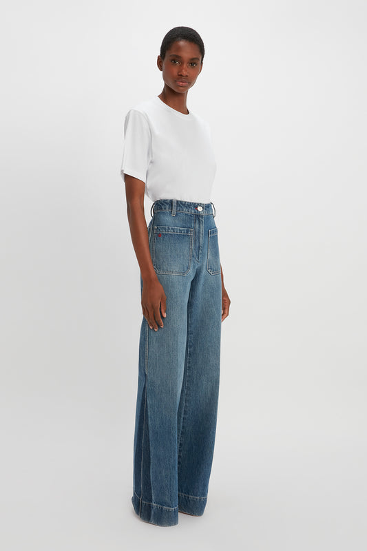 Person standing against a plain background, wearing a white short-sleeve shirt and high-waisted, 70s-inspired Alina High Waisted Jean In Shadow Wash by Victoria Beckham.